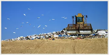 A bulldozer compacts a large pile of waste at a landfill site under a clear blue sky, with birds flying overhead.